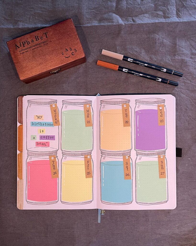 Colorful weekly planner with jar designs, markers, and wooden alphabet box on a textured fabric background.