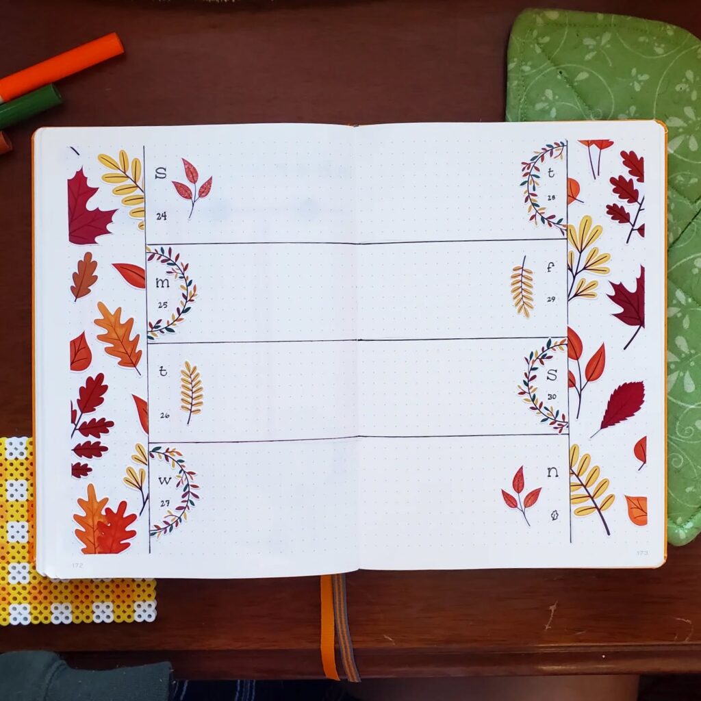 Open bullet journal with colorful autumn leaf illustrations, featuring weekly planner layout on a wooden table.