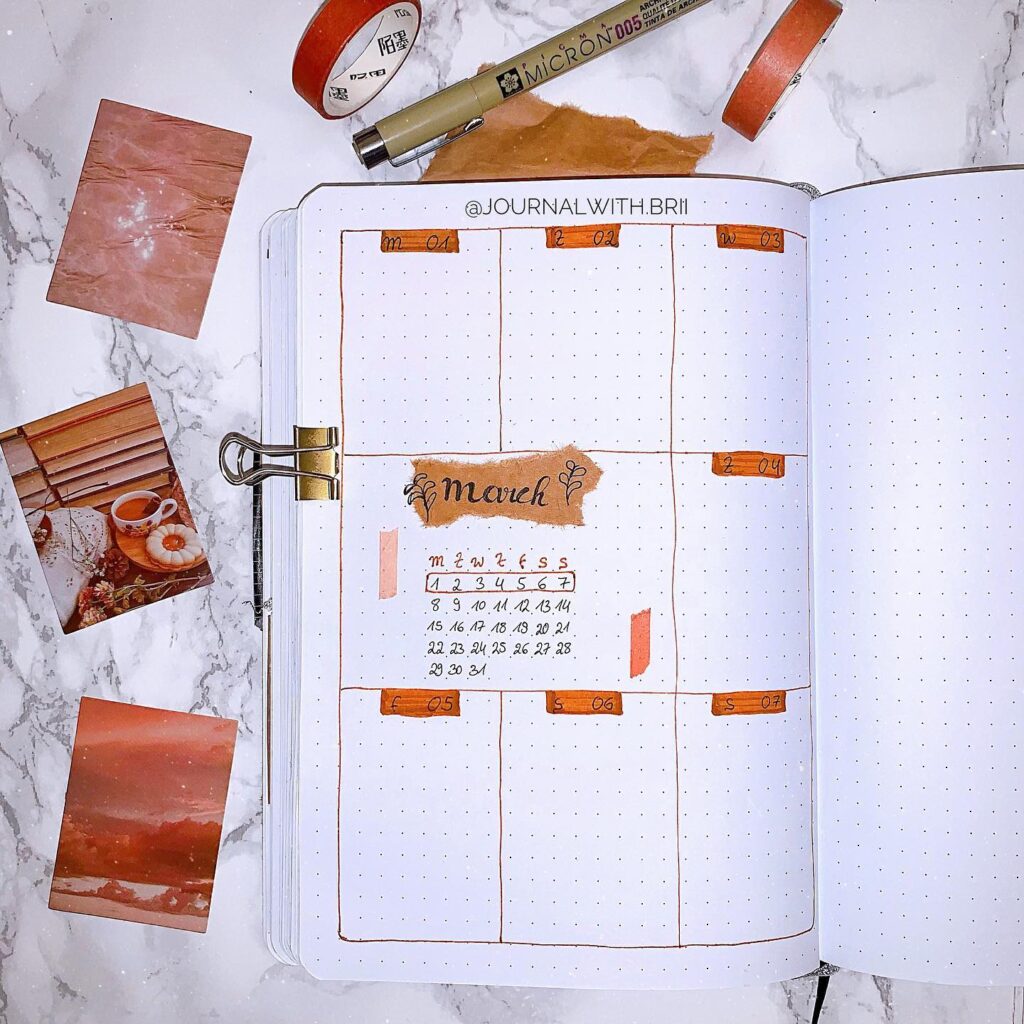 Bullet journal for March with orange accents, pen, and photos nearby on a marble surface.