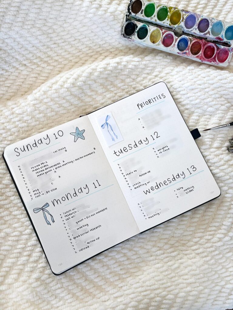 Open bullet journal with weekly spread and watercolor set on cozy white blanket.