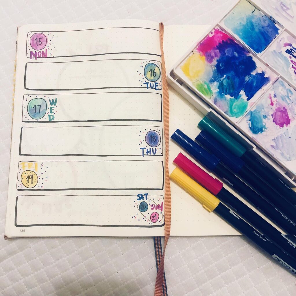 Bullet journal weekly planner with colorful pens and paint palette on a white surface.
