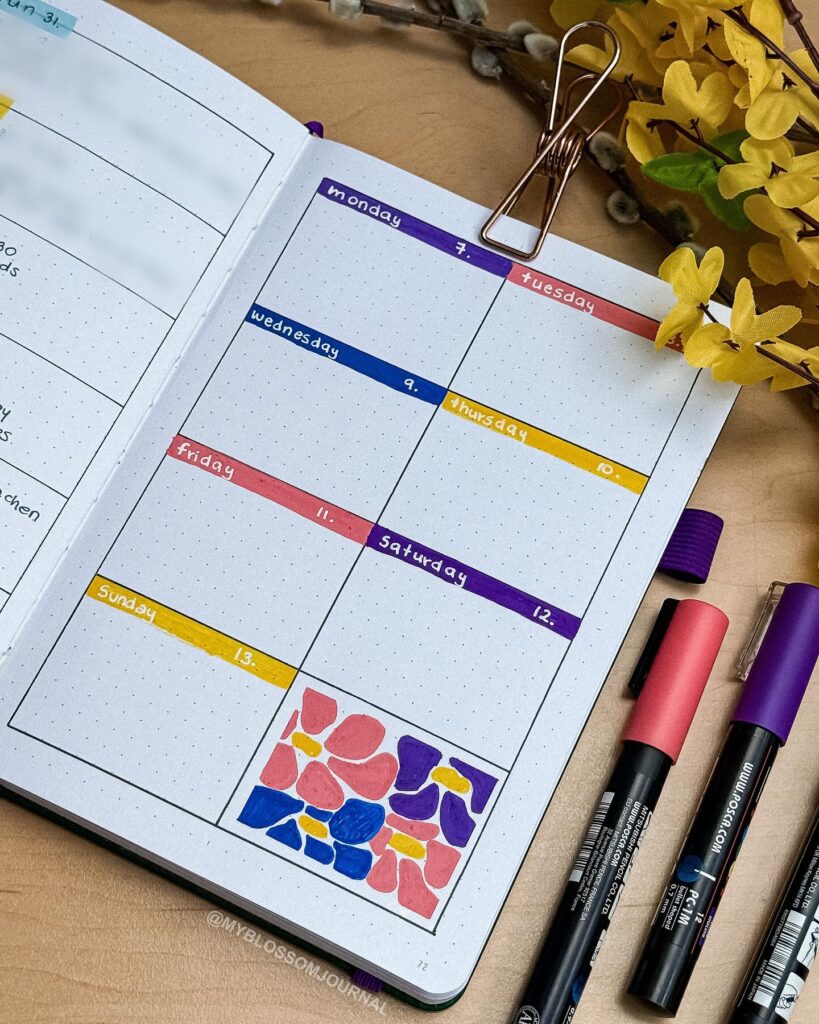 Bullet journal layout with color-coded days, markers, and floral doodle on a table with yellow flowers.