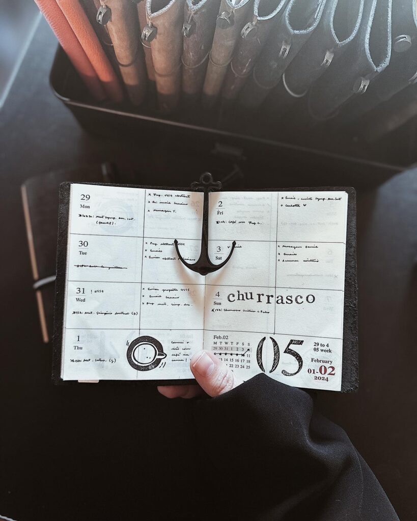 Hand holding an open planner with weekly schedule, surrounded by pens and office supplies.