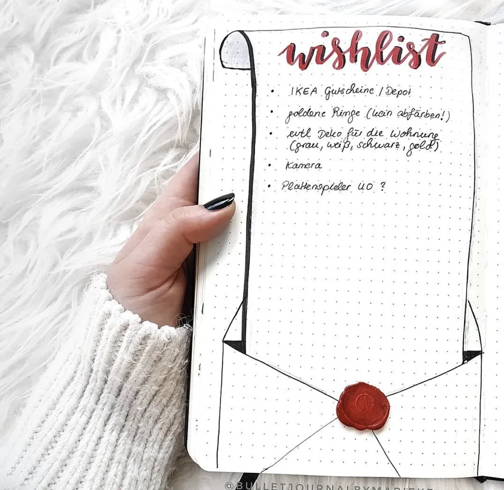 Hand holding a bullet journal with wishlist, decorative envelope drawing, and cozy white sweater sleeve.