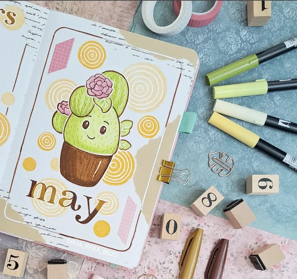 Cute cactus drawing in a May bullet journal surrounded by pens, stamps, and washi tape for crafting inspiration.