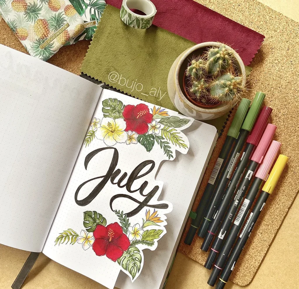 July bullet journal with floral design, colorful markers, cactus plant, and tropical-themed decor.