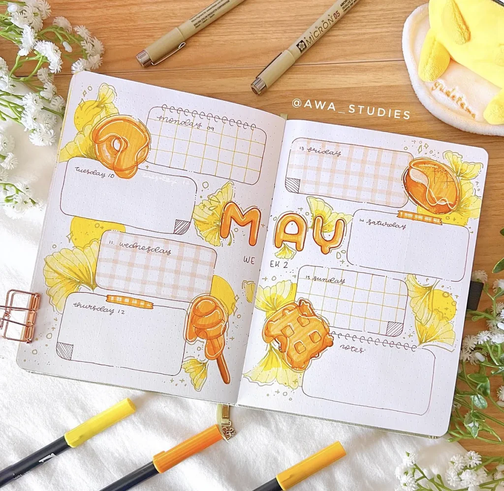 Colorful May bullet journal spread with flowers, honeycomb theme, and handwriting surrounded by pens on a wooden desk.