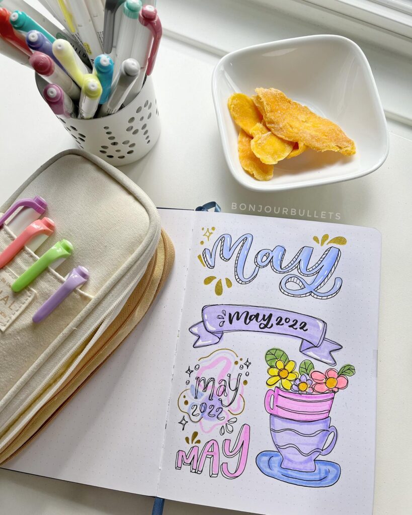 Creative May bullet journal with colorful pens, pencil case, and dried mango slices on a desk.