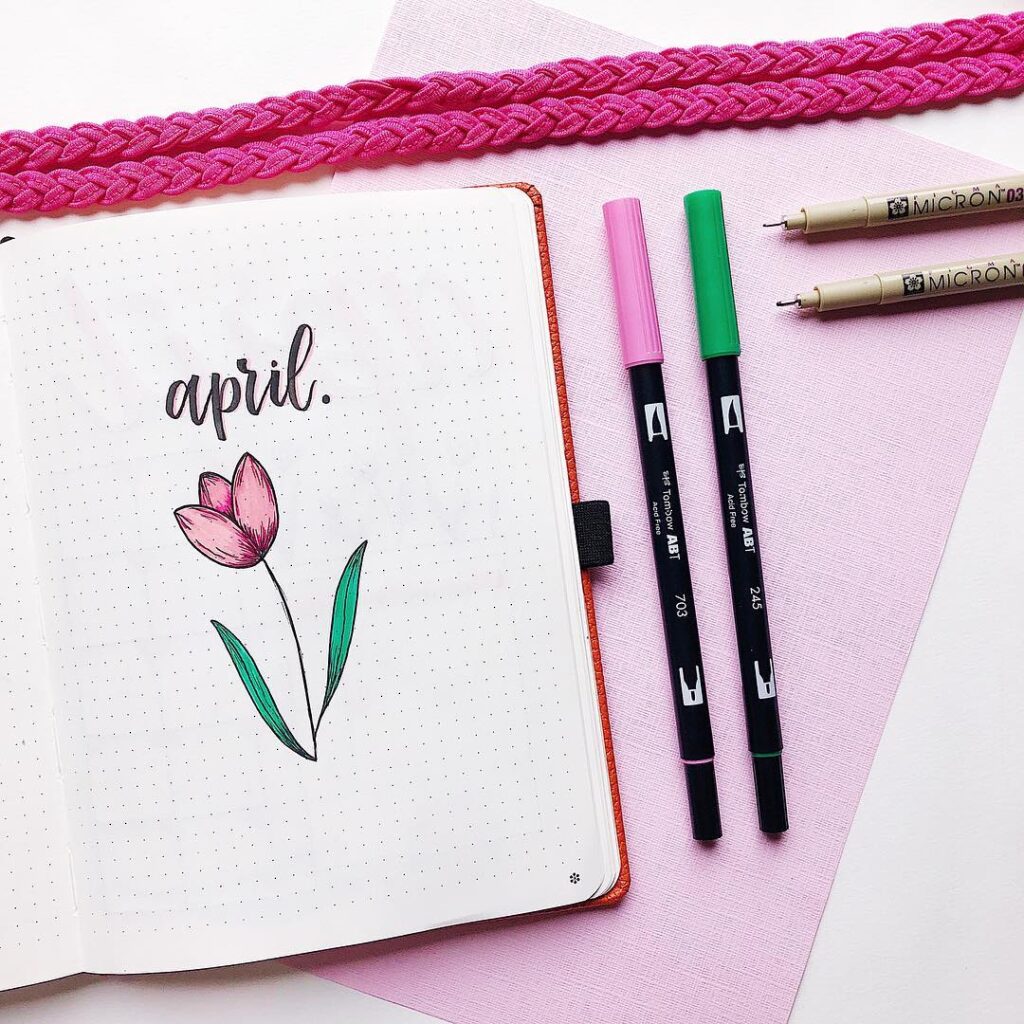 Bullet journal open to April with flower doodle, surrounded by pens and pink braided decor on a dotted page.