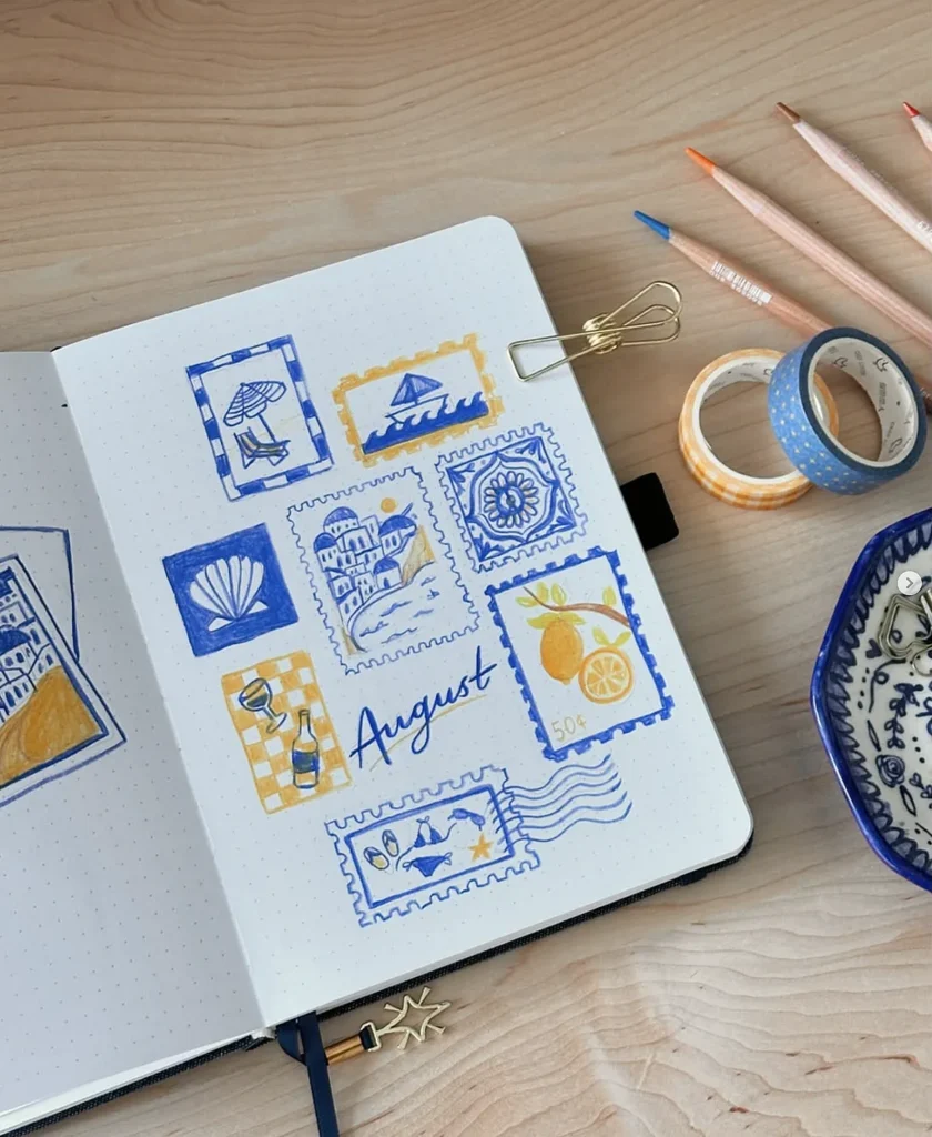 Notebook page with artistic stamps and the word August, surrounded by colored pencils and tape on a wooden desk.