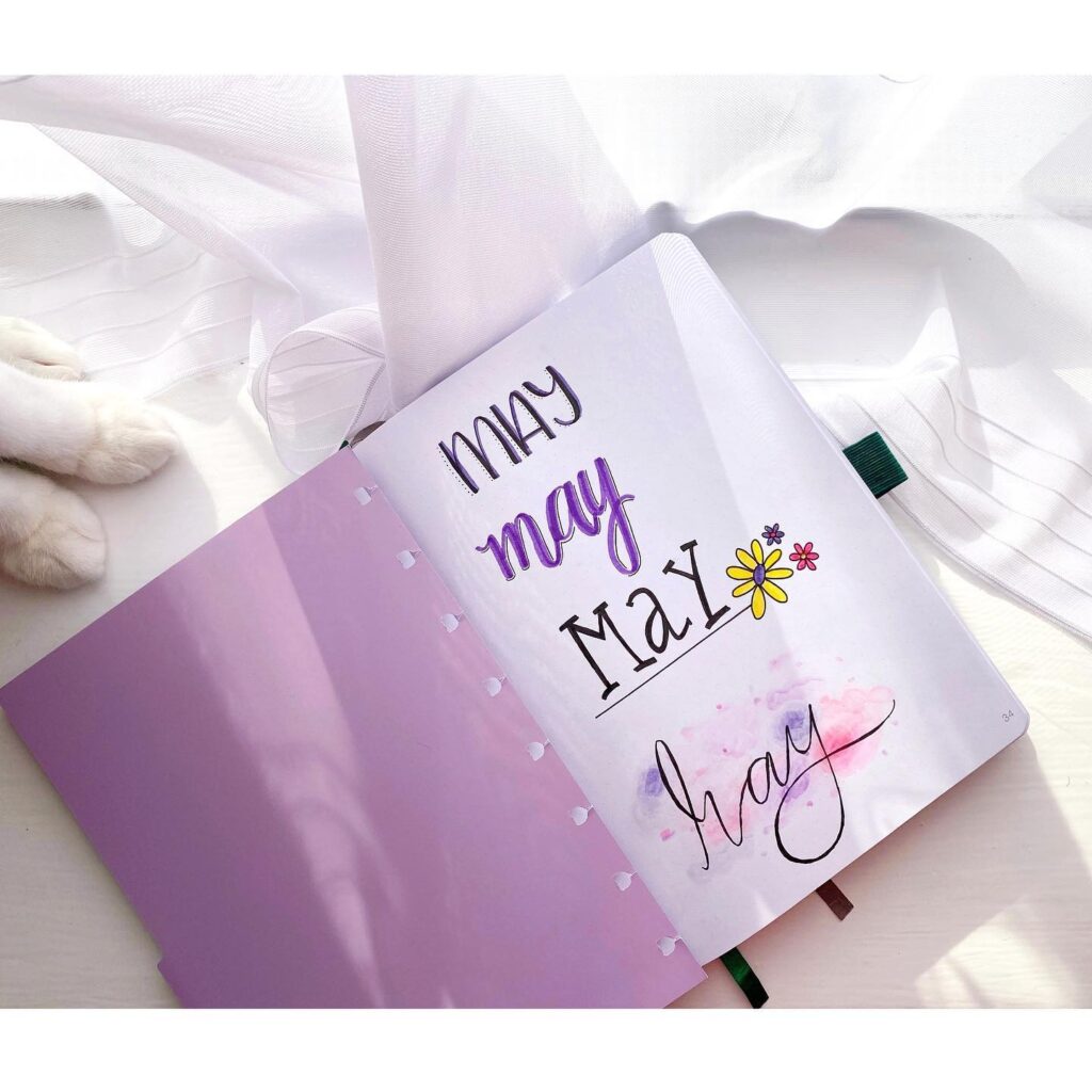 Open planner displaying artistic May lettering, adorned with flowers, on a sunny windowsill with sheer curtains.