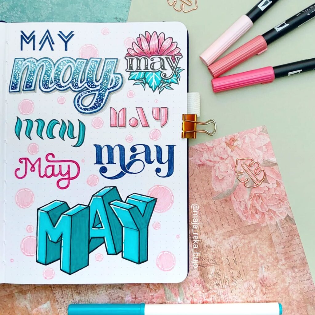 Creative May lettering designs in various styles and colors on paper with markers and floral accents.