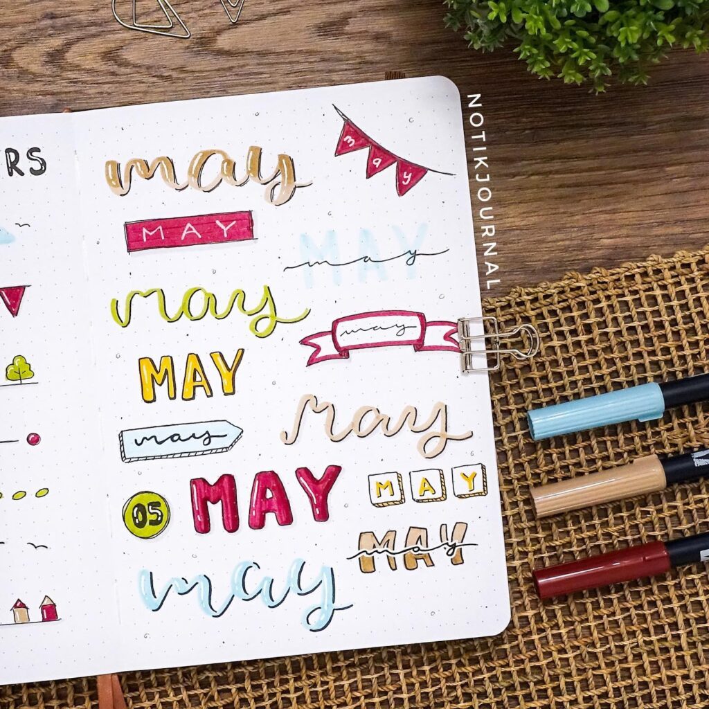 Notebook page with creative May lettering in various styles, surrounded by pens and paper clips on a wooden surface.
