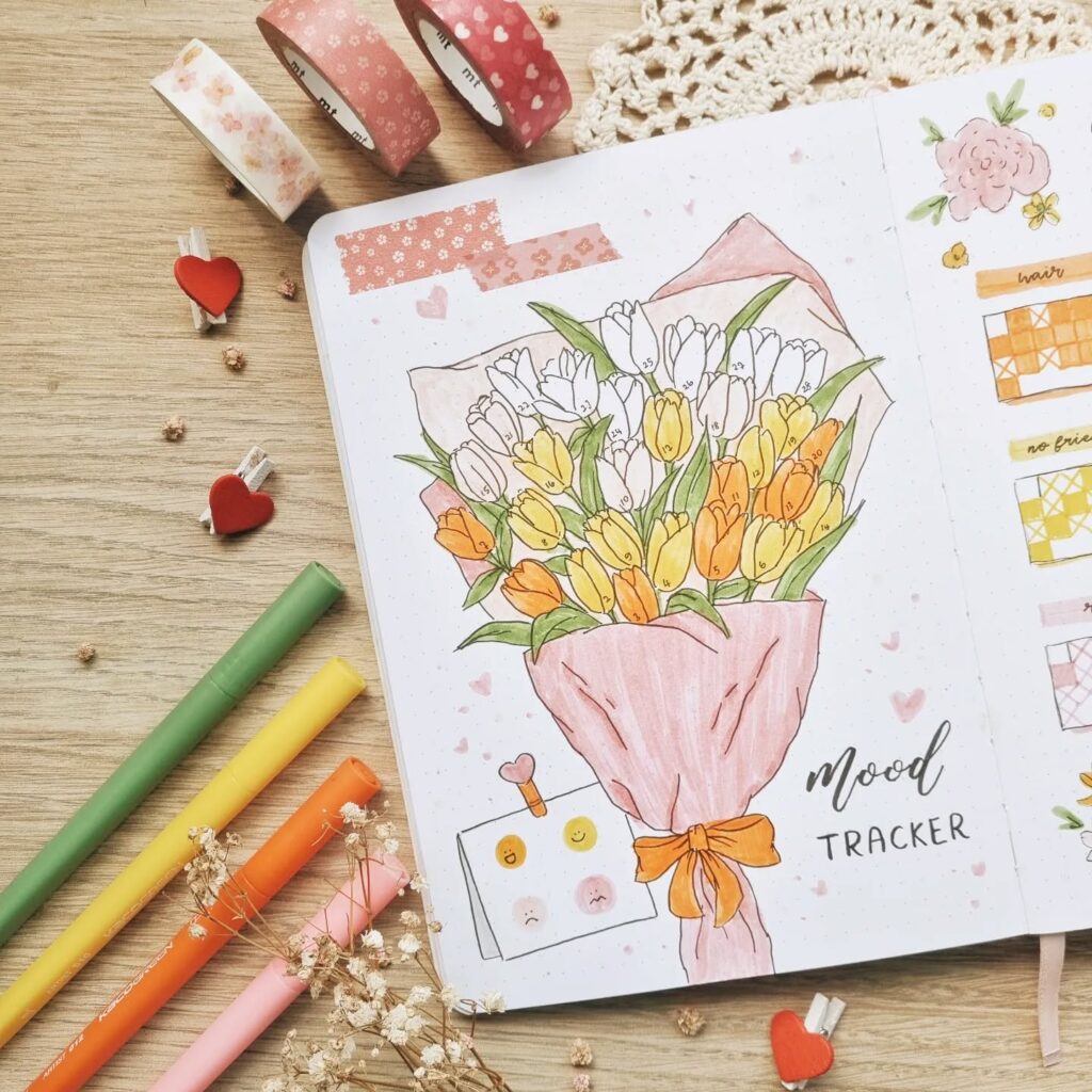 Bullet journal with tulip bouquet illustration, mood tracker, colorful pens, and washi tape on a wooden surface.
