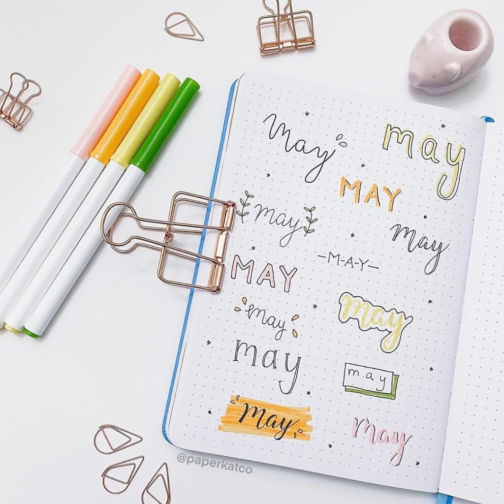 Bullet journal with various May lettering styles, surrounded by pens and clips. Creative monthly planner design.