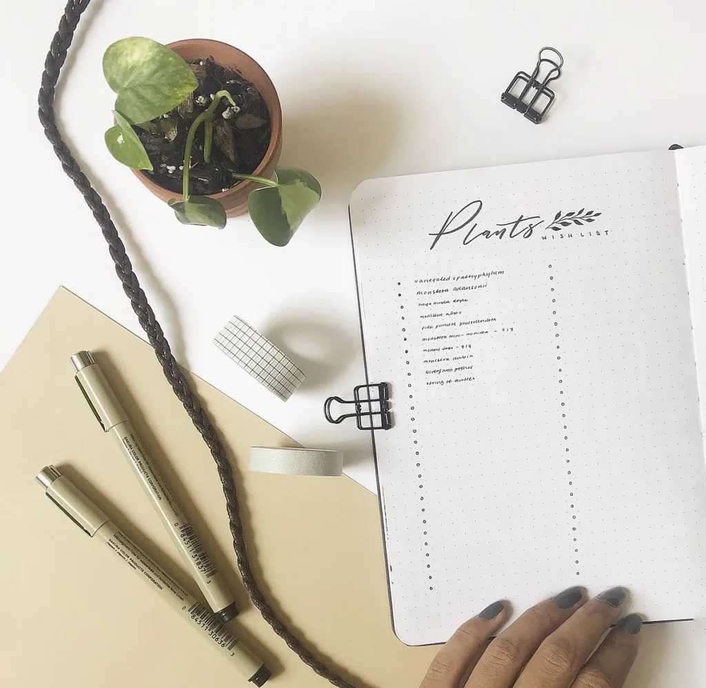 Open journal with plant wishlist, beside potted plant, pens, and paper clips on a white desk.
