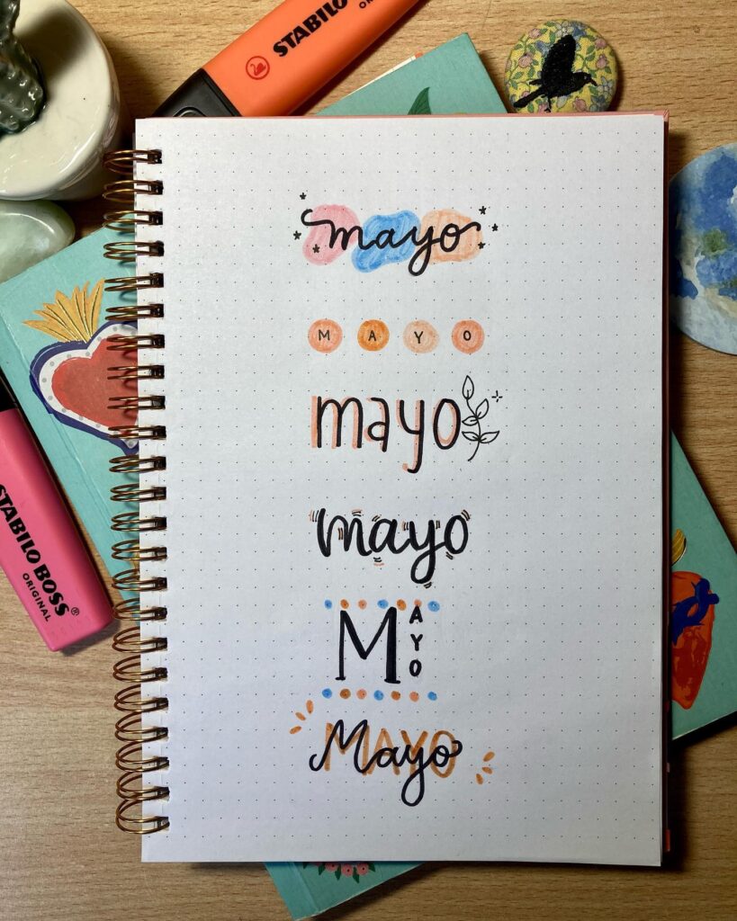 Notebook page with creative hand-lettering for Mayo in various styles, surrounded by colorful markers and notebooks.