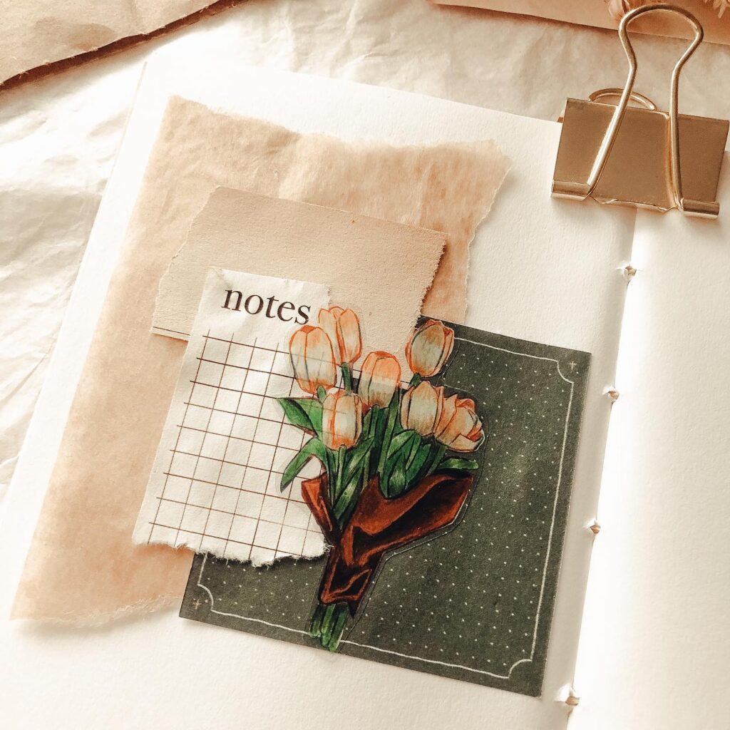 Journal page with floral illustration, scattered paper layers, and notes label, clipped with a gold binder.