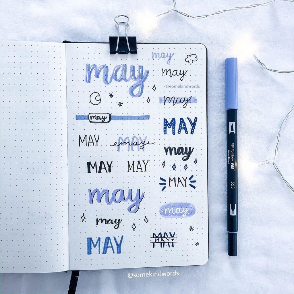 Journal page displaying various hand-lettered May designs in blue and black with a marker beside it.