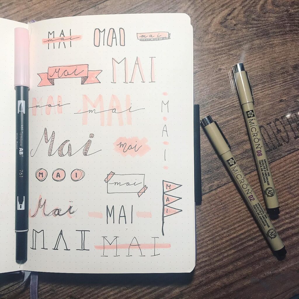 Notebook with various Mai lettering styles in pink and black, surrounded by pens on a wooden surface.