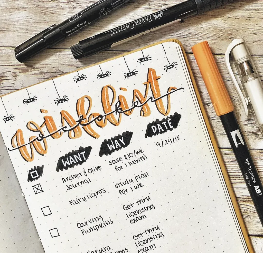 Bullet journal wishlist with pens and markers on a wooden table, featuring doodles of spiders and notes.