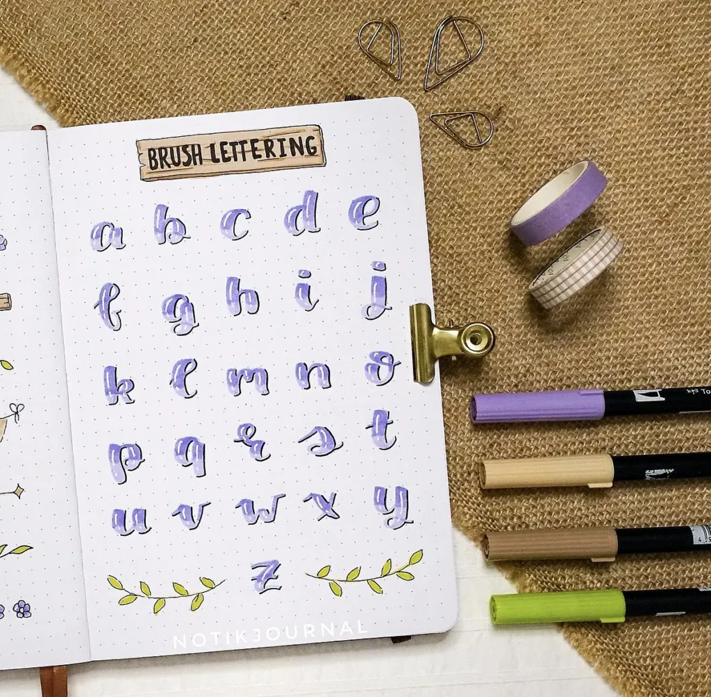 Calligraphy alphabet guide in notebook with brush pens, decorative tape, and paper clips on a burlap background.