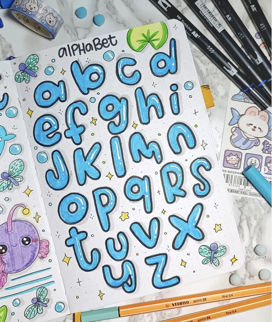 Colorful hand-drawn lowercase alphabet in a sketchbook with cute doodles and art supplies surrounding it.
