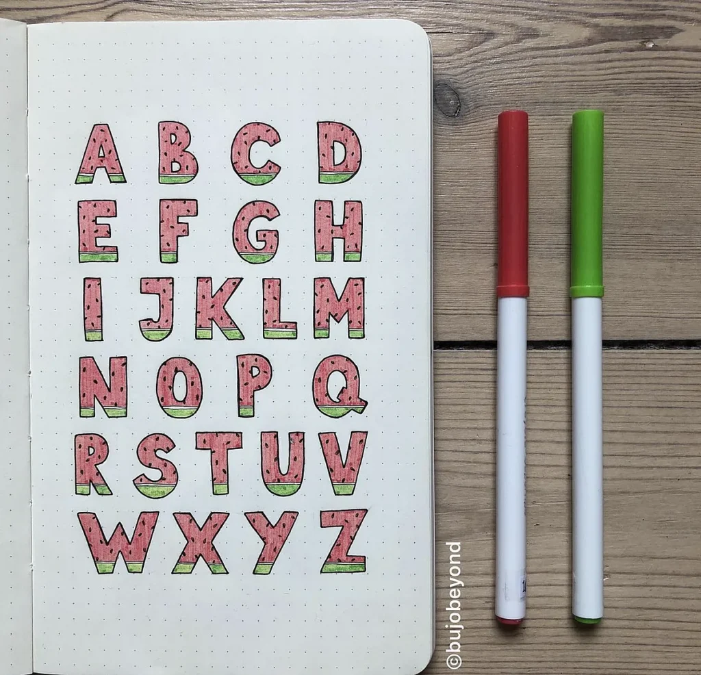 Alphabet letters styled as watermelons with red and green pens on a wooden table background.