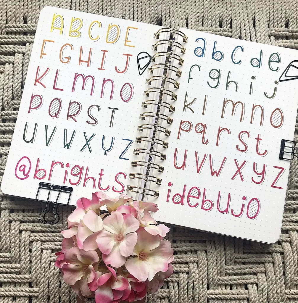 Creative bullet journal with colorful alphabet letters, open on a knitted rug, featuring a pink flower.