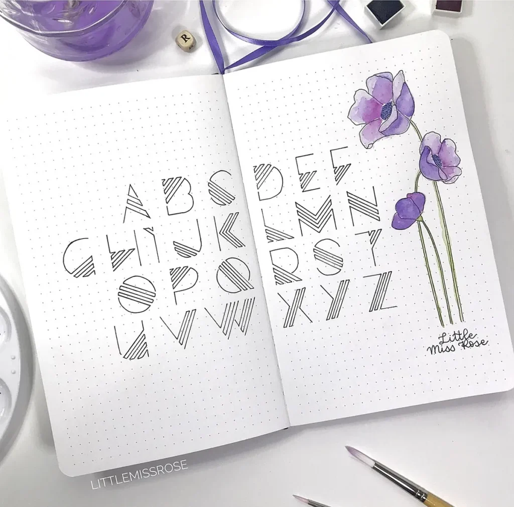 Bullet journal open to a page with artistic alphabet design and watercolor purple flowers.