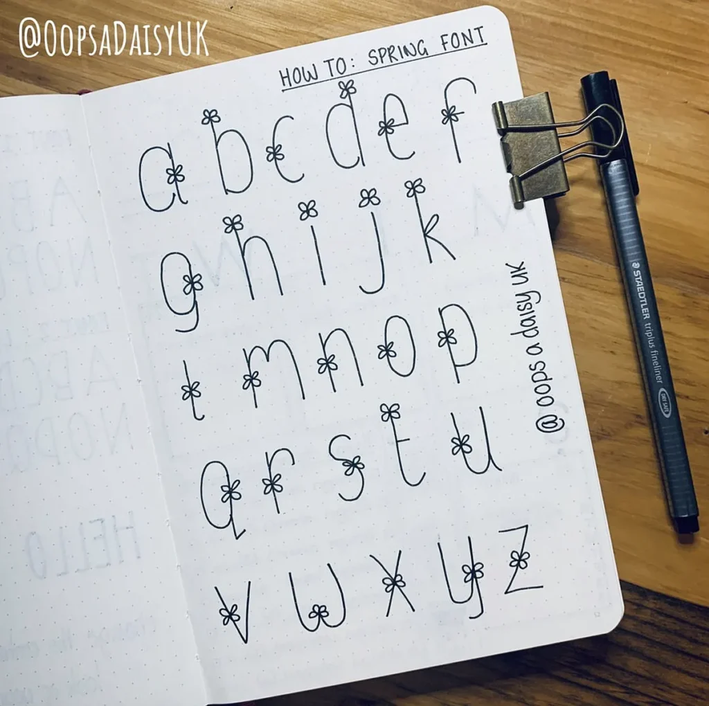 Alphabet in handwritten spring font with floral details, notebook and pen on wooden table.