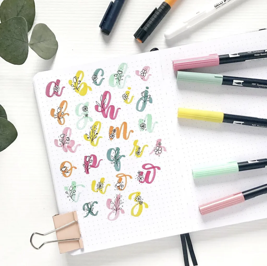 Colorful hand-lettered alphabet in a notebook, surrounded by markers on a white surface.