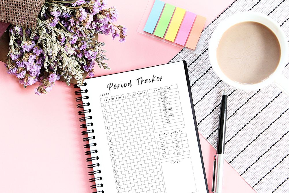 Period tracker on pink background with coffee, pen, sticky notes, and flowers. Organizational tool for menstrual health.