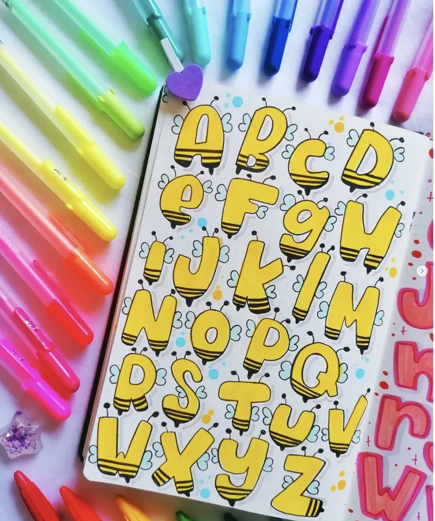Colorful markers surround a notebook with bee-themed alphabet letters in yellow, enhancing creative lettering.