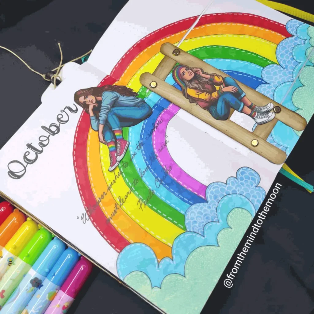 Colorful October journal page with rainbow, clouds, and girl illustration. Markers displayed beside the open notebook.