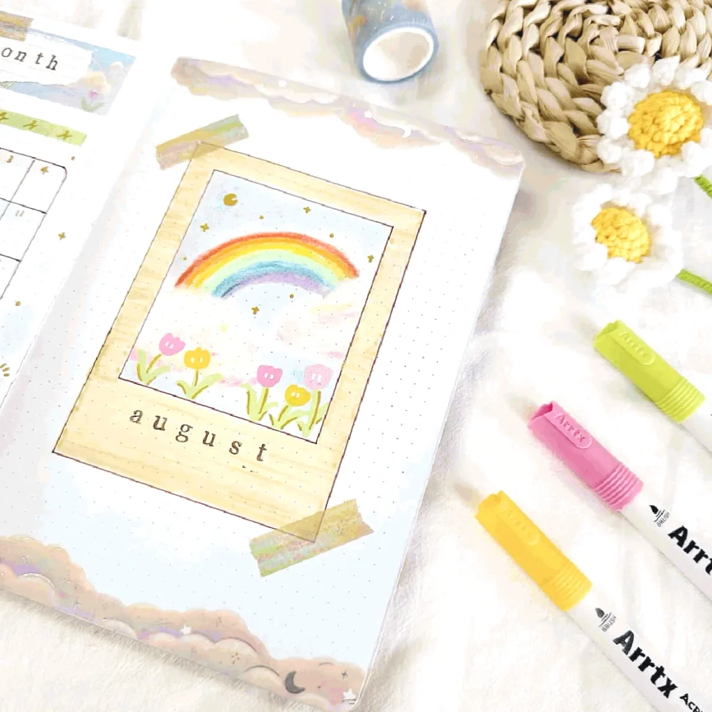 August bullet journal page with rainbow, stars, and flowers, surrounded by colorful markers and decor items.