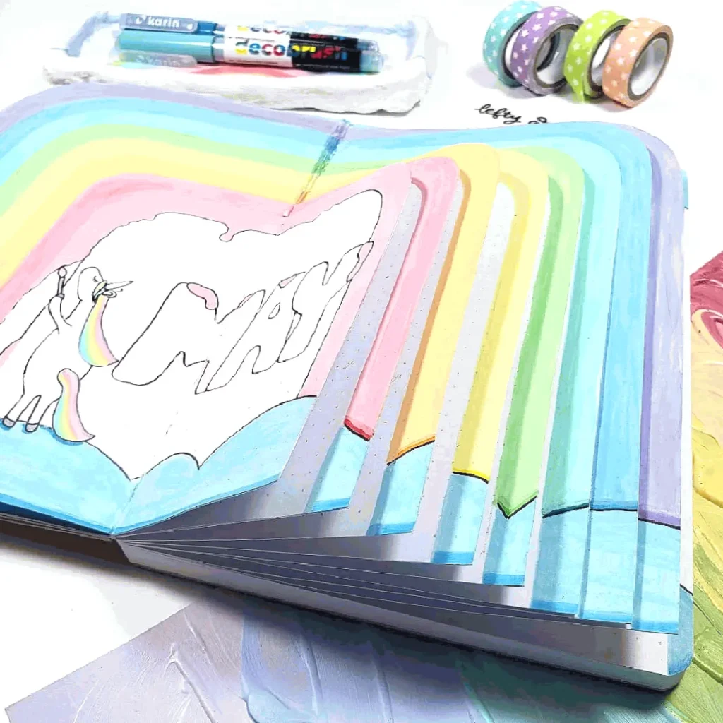 Colorful notebook with rainbow pages, showcasing the word MAY and whimsical designs, surrounded by art supplies.