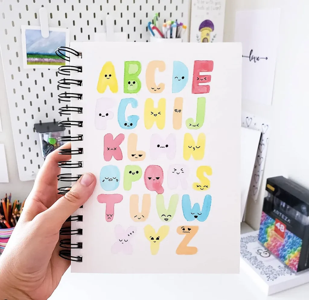 Colorful alphabet art with cute faces on letters in a spiral notebook held in hand against a creative desk background.