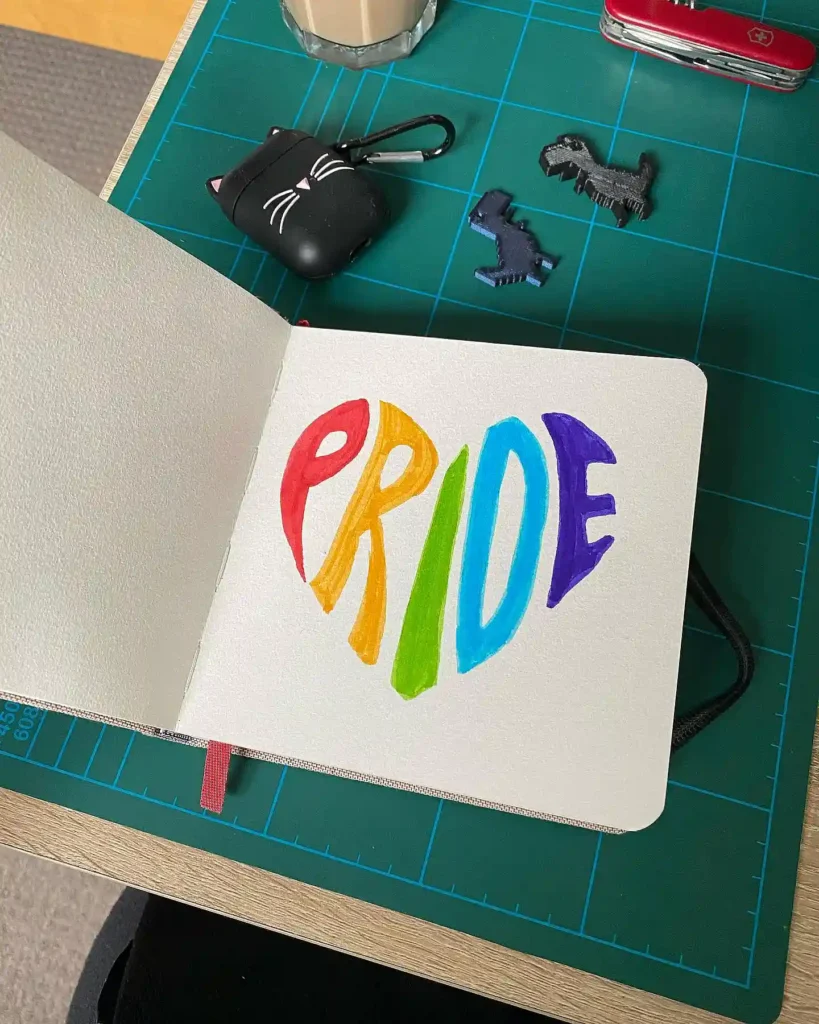 Notebook with colorful Pride drawing on desk, surrounded by coffee, cat-shaped keychain, and small toys.