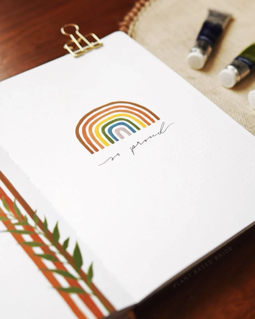 Open notebook with rainbow design and so proud text, featuring a gold clip and paint tubes on a wooden surface.