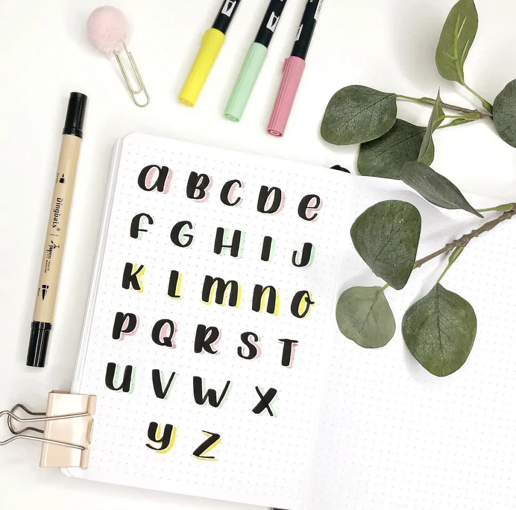 Alphabet calligraphy practice in journal with colorful pens and green leaves on a white background.