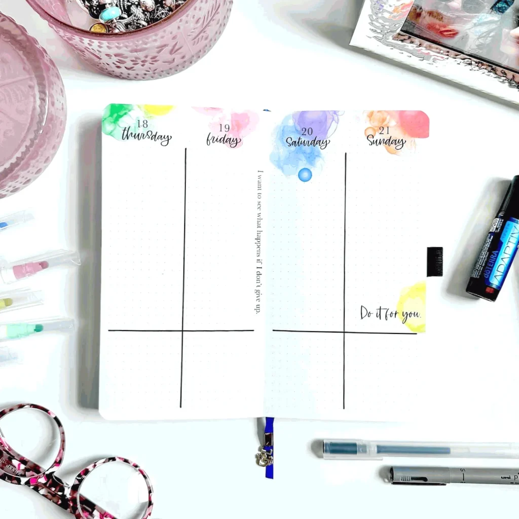 Open planner with colorful watercolor headings, surrounded by stationery on a white desk.