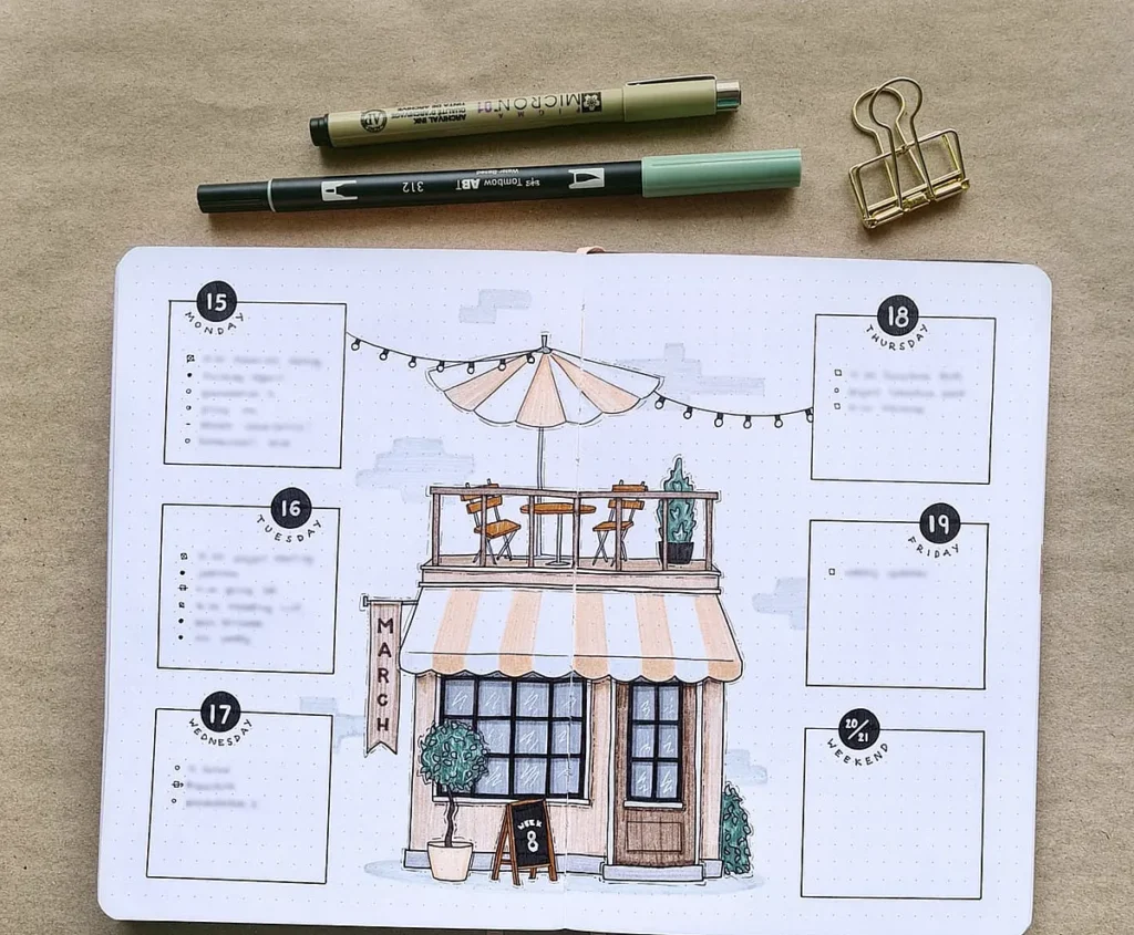 Bullet journal open to a March weekly layout with a café doodle, surrounded by pens and a paper clip.