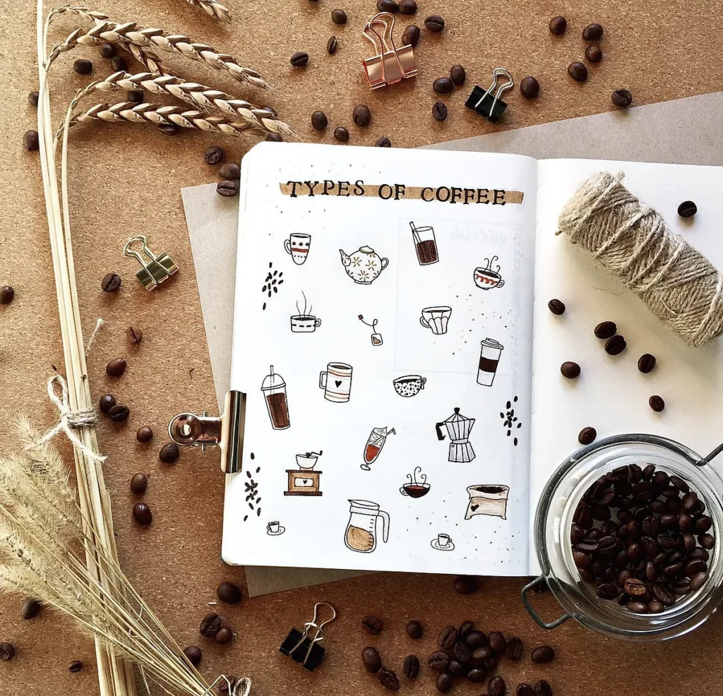 Illustrated coffee types in a journal surrounded by coffee beans, wheat, twine, and paper clips on a rustic background.
