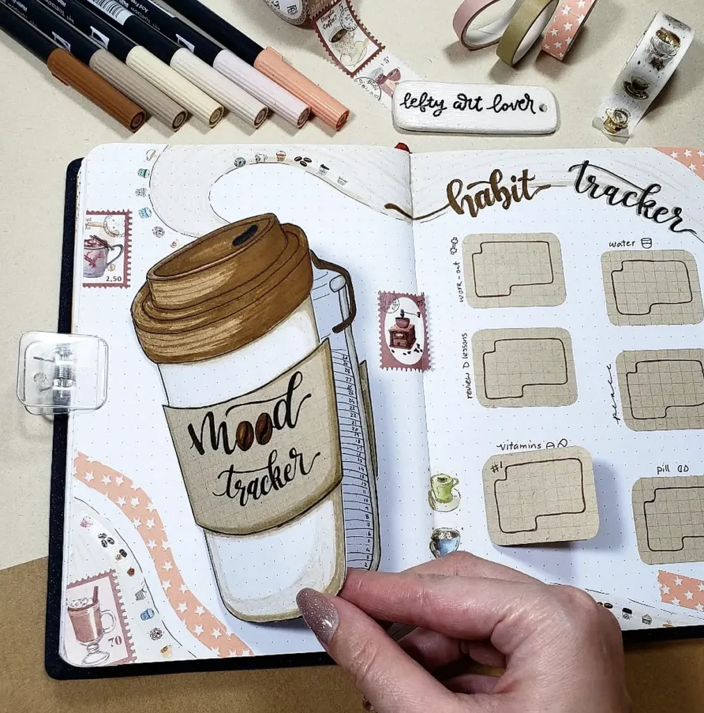 Bullet journal with mood and habit trackers, coffee cup illustration, and various markers and washi tape.