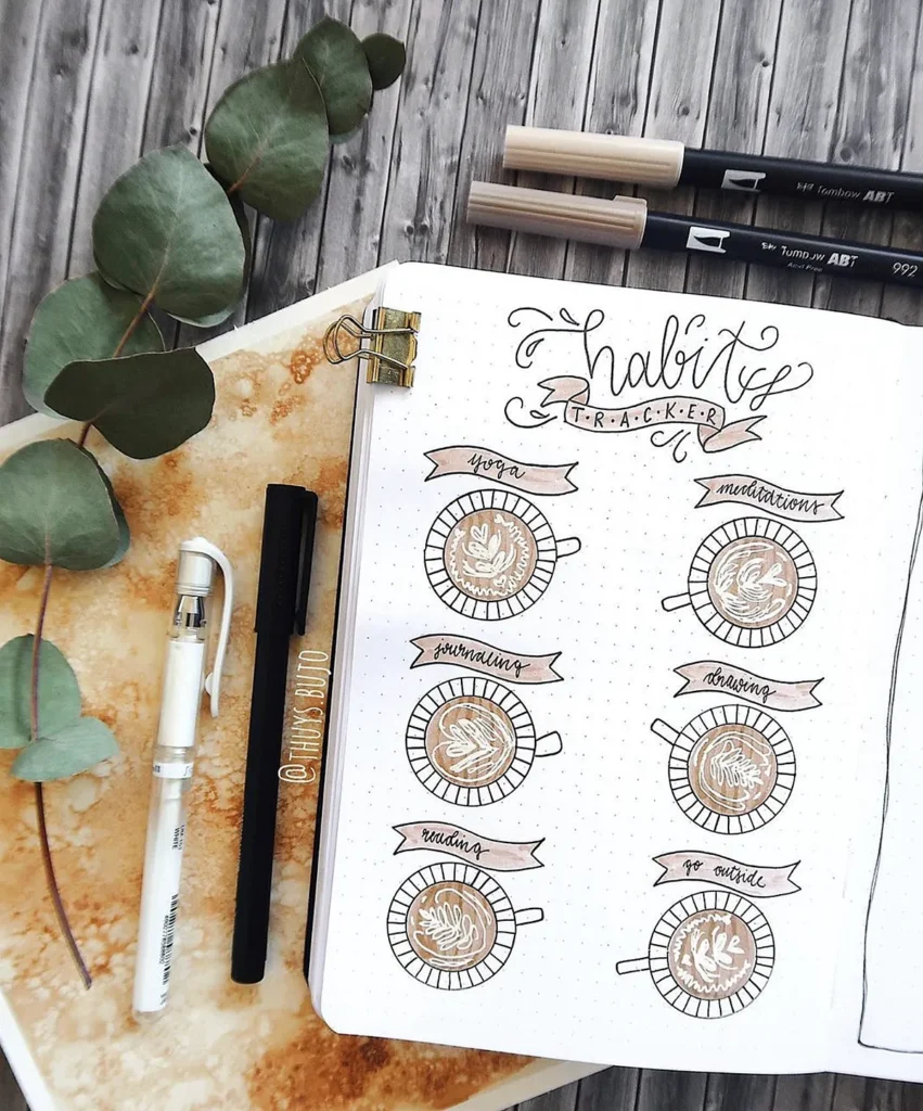 Bullet journal habit tracker with coffee-themed doodles, pens, and decorative leaves on a wooden table.
