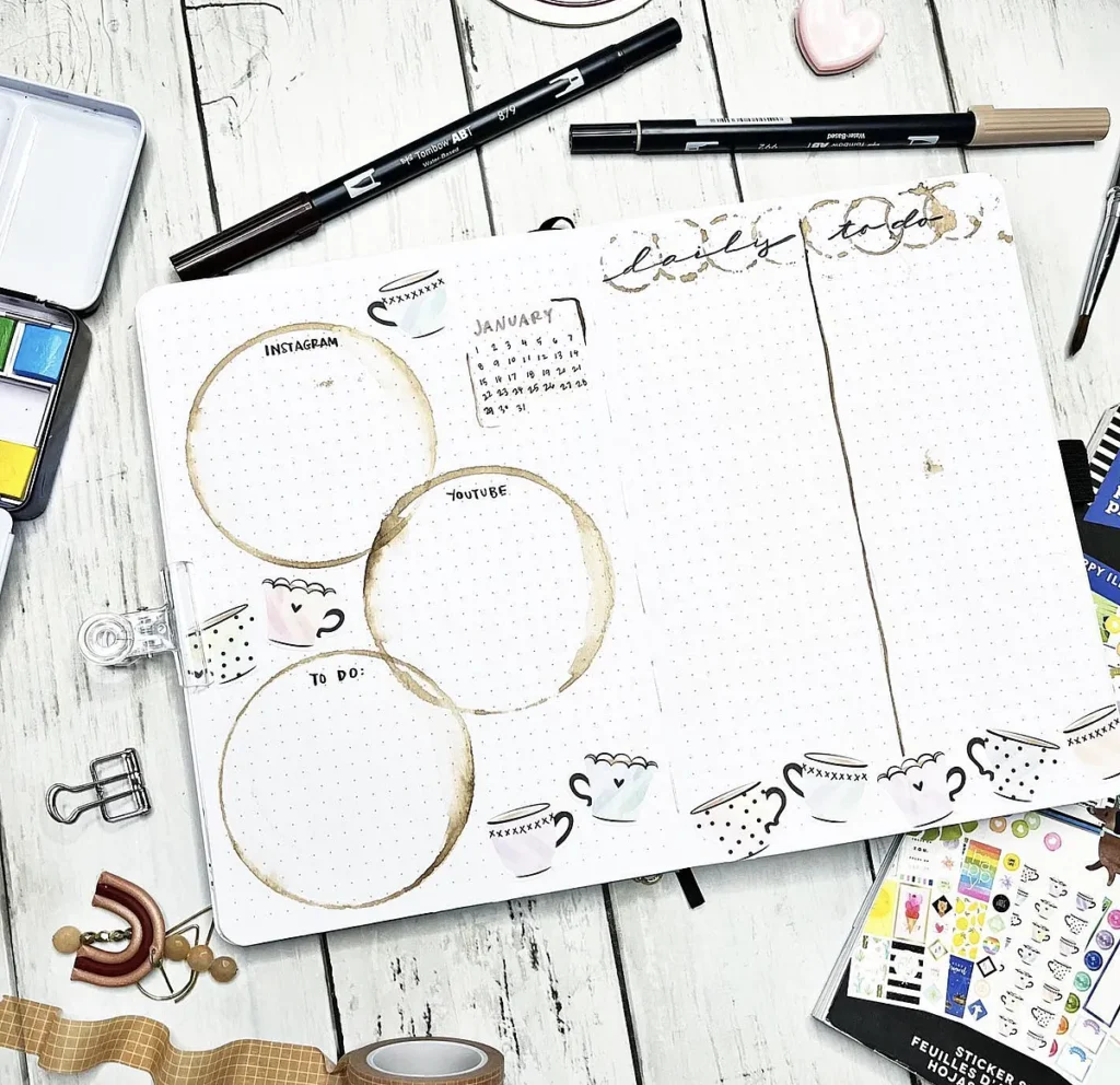 Open bullet journal on wooden table with pens, stickers, and coffee cup designs for organizing tasks and plans.