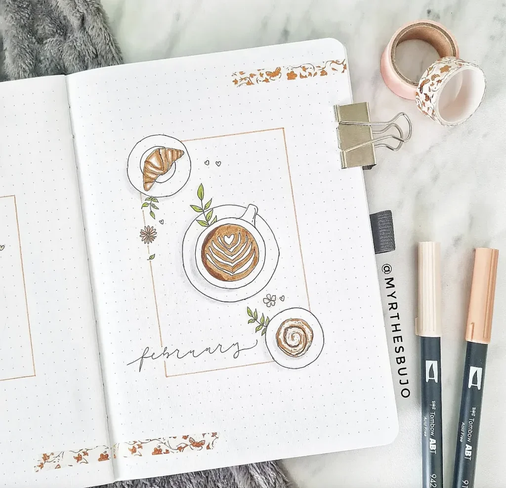 Bullet journal page with coffee, croissant, and cinnamon roll illustrations, labeled February. Art supplies nearby.