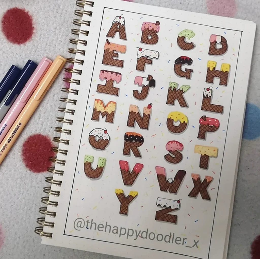 Alphabet with ice cream cone designs on a spiral notebook page, surrounded by pens on a polka dot background.