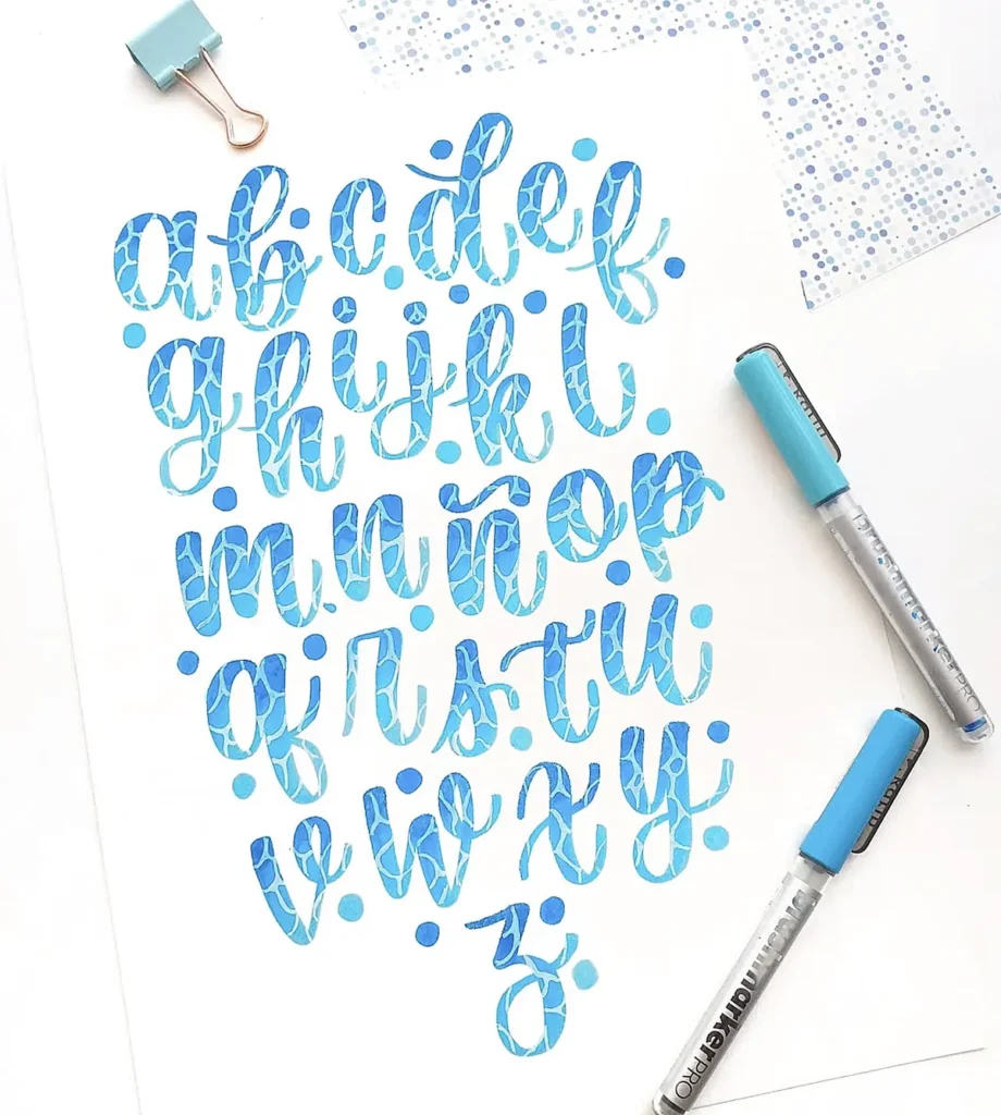Blue watercolor calligraphy alphabet artwork with pens on white paper.
